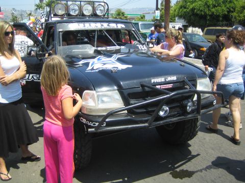 1990 Ford Bronco - black Ford Ranger prerunner with a roof light bar, star graphic and Free JMS decal is surrounded by spectators on a street.