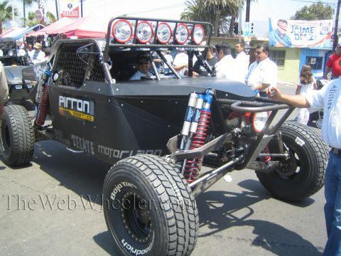 1990 Ford Bronco - black Pirron tube-chassis off-road buggy with a roof light bar and BFGoodrich tires waits on a street lined with Tecate banners.