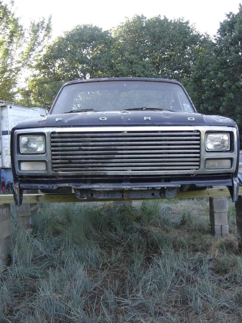 Front clip of a faded black 1980s Ford truck cab raised on blocks outdoors