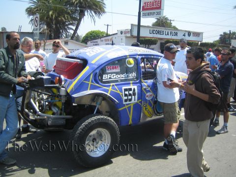 1990 Ford Bronco - blue and yellow Baja-prepped VW Beetle number 554 with BFGoodrich and SCORE decals is surrounded by people on a street.