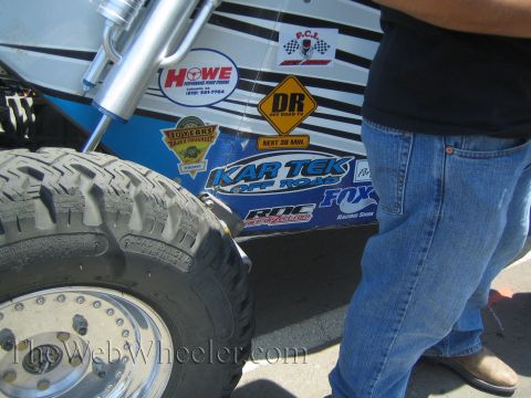 1990 Ford Bronco - Close-up of an off-road race car's rear tire and silver coilover shock beside Kartek and Fox sponsor decals, with a man standing by.