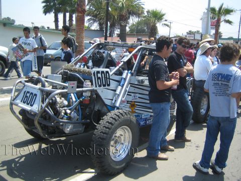 1990 Ford Bronco - silver and black number 1600 mid-engine off-road race buggy sits on a palm-lined street surrounded by spectators.