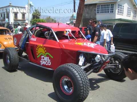 1990 Ford Bronco - red off-road race buggy with Del Taco and Coca-Cola branding sits on a palm-lined street as spectators gather around it.