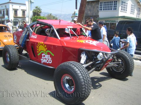 1990 Ford Bronco - red Del Taco off-road race buggy with Coca-Cola decals is inspected by onlookers on a sunny palm-lined street.