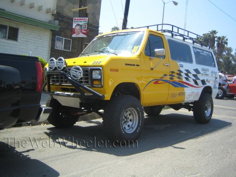 1990 Ford Bronco - yellow lifted Ford support van number 222 with a roof rack, front light pods and checkered graphics sits on a street.