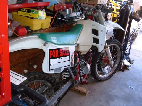 1985 Ford Bronco II - A dirt bike with a "FOR SALE" sign is parked in a garage, surrounded by tools and supplies.