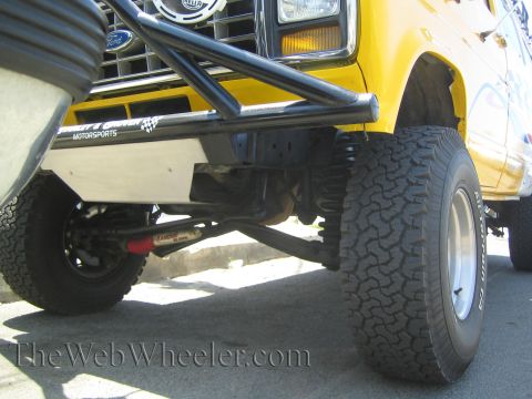 1990 Ford Bronco - Close-up of the undercarriage of a lifted yellow Ford with a Ford-badged bumper, exposed front suspension, shock and a BFGoodrich tire.