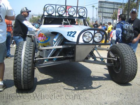 1990 Ford Bronco - silver tube-chassis off-road racing buggy numbered 122 with a roof-mounted light bar and twin round headlights, people standing alongside.