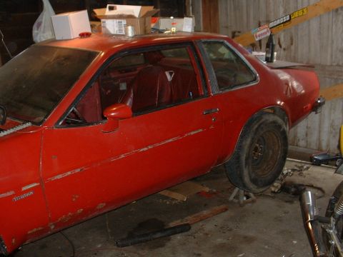 1980 Chevrolet Monza - A red coupe, possibly a 1975 model, sits on jack stands in a garage with visible wear and tear.