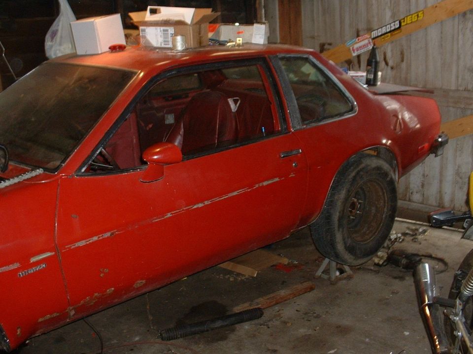 Faded red Chevrolet Monza coupe on a jack stand in a dusty wooden garage