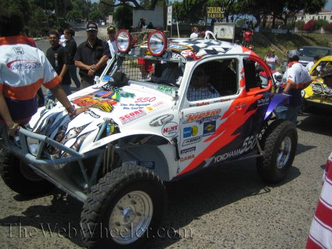 1990 Ford Bronco - white and orange VW Beetle-based Baja race car numbered 550 with a checkered-flag roof and many sponsor decals, crew standing around it.