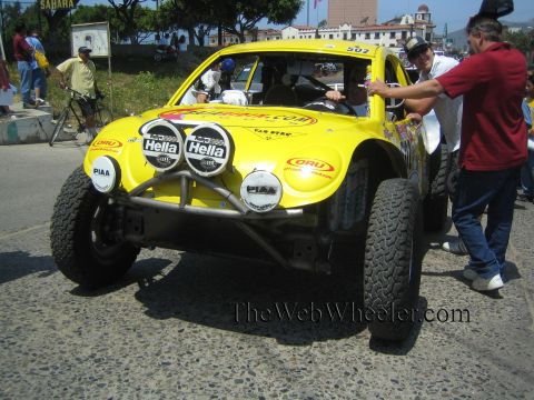1990 Ford Bronco - Front view of a yellow VW Beetle-based Baja race car numbered 502 with twin round driving lights and Hella stickers, a man reaching toward…