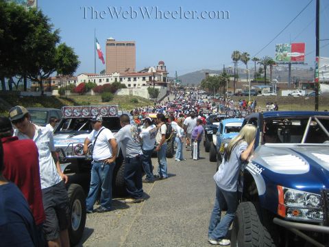 1990 Ford Bronco - crowded sunny street packed with spectators and off-road race trucks, a large Mexican flag and a tall hotel building in the background.