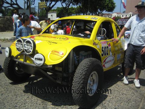 1990 Ford Bronco - yellow VW Beetle-based Baja race car numbered 502 with twin round lights and Hella decals parked on a street, a man standing beside it.