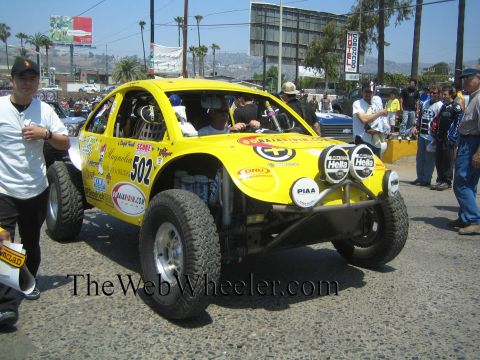1990 Ford Bronco - Three-quarter front view of a yellow VW Beetle-based Baja race car numbered 502 covered in sponsor decals, spectators and palm trees behind.