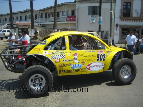 1990 Ford Bronco - Side view of a yellow VW Beetle-based Baja race car numbered 502 with BFGoodrich and Magnolia Gowns decals, parked before storefronts.