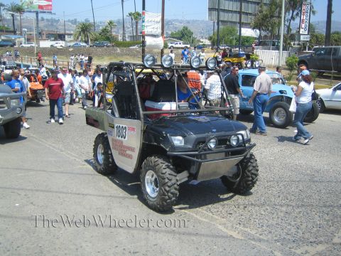 1990 Ford Bronco - dark off-road UTV side-by-side numbered 1803 with a roof light bar driving past spectators on a sunny street.