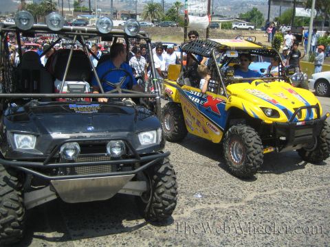 1990 Ford Bronco - black Polaris off-road UTV and a yellow side-by-side, both with roof light bars, parked among spectators on a sunny street.