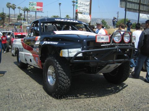 1990 Ford Bronco - black and red off-road race truck with a tube front bumper, dual round lights and long-travel front suspension, spectators around it.