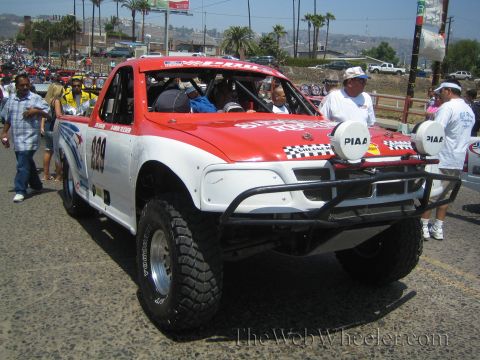 1990 Ford Bronco - Front three-quarter view of a red and white off-road race truck numbered 999 with twin round PIAA lights and a tube bumper, crowd around it.