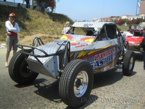 1990 Ford Bronco - silver tube-frame off-road buggy with Laminas Fastele graphics parked beside a yellow buggy numbered 1601, a woman standing at left.