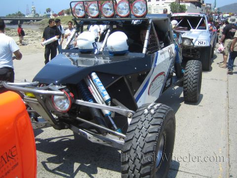 1990 Ford Bronco - silver and blue tube-frame off-road buggy with a roof light bar and crash helmets inside, parked on a seaside ramp with other race cars.