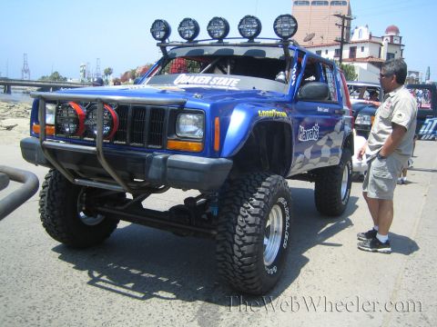 1990 Ford Bronco - lifted blue Jeep Cherokee with a roof-mounted light bar, tube front bumper and chunky off-road tires parked near a waterfront, a man beside…