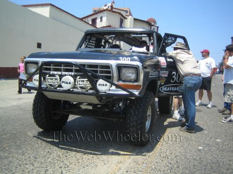 1990 Ford Bronco - black vintage Ford pickup desert race truck number 300 with roof-mounted lights and Hella/PIAA driving lamps on pavement.