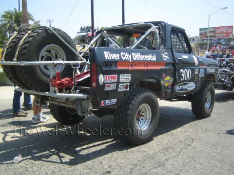1990 Ford Bronco - Rear three-quarter view of a black Ford desert race truck number 300 carrying two spare tires on a red rack in the bed.