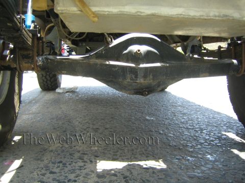 1990 Ford Bronco - Underside rear view of a truck showing the black differential housing, axle tubes and leaf-spring suspension over pavement.