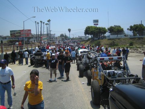 1990 Ford Bronco - long street packed with off-road race vehicles staged in a row while crowds of spectators gather under a bright sky.