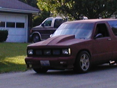 1998 Ford Taurus - A maroon pickup truck with a custom hood scoop and aftermarket wheels is parked on a street in front of a garage.