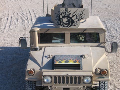 1979 Ford Bronco - A tan military vehicle is shown from the front, a skull and flames decal on its roof-mounted gun shield and hood.