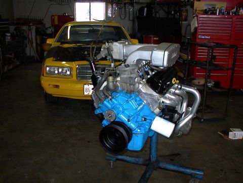 1986 Mercury Cougar - A yellow car sits in a garage with its hood open, while a separate engine is displayed on a stand in the foreground.
