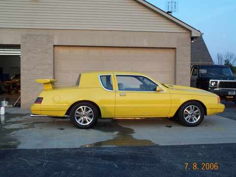 1986 Mercury Cougar - A yellow 1986 Mercury Cougar LS coupe is parked next to a black pickup truck in front of a garage.
