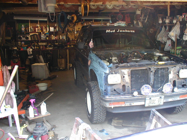Lifted dark SUV mid-build inside a cluttered garage, engine bay open, 'Mud Jamboree' on rear glass