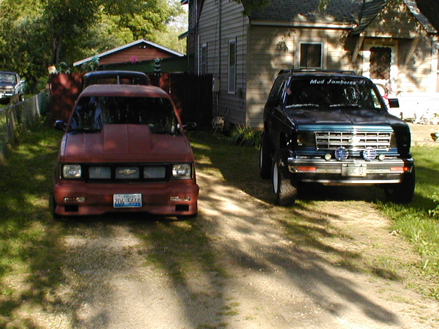 Lowered brown S-10 and a lifted dark Chevy SUV parked in a yard between houses