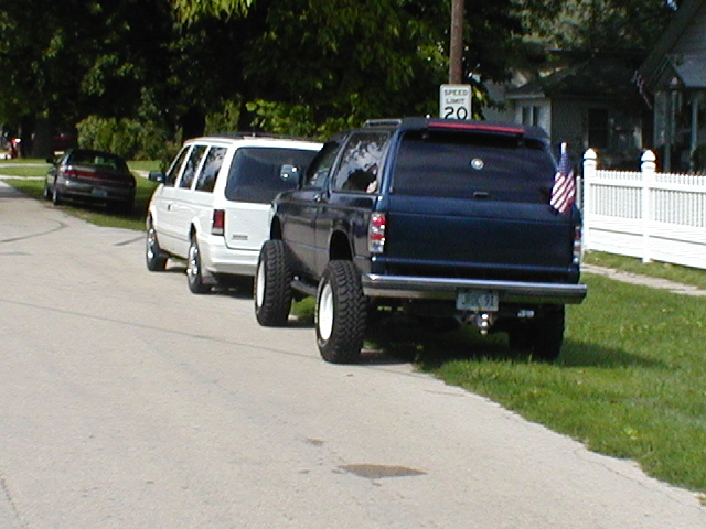 Lifted dark blue pickup truck parked behind white minivan on a street. American flag on truck