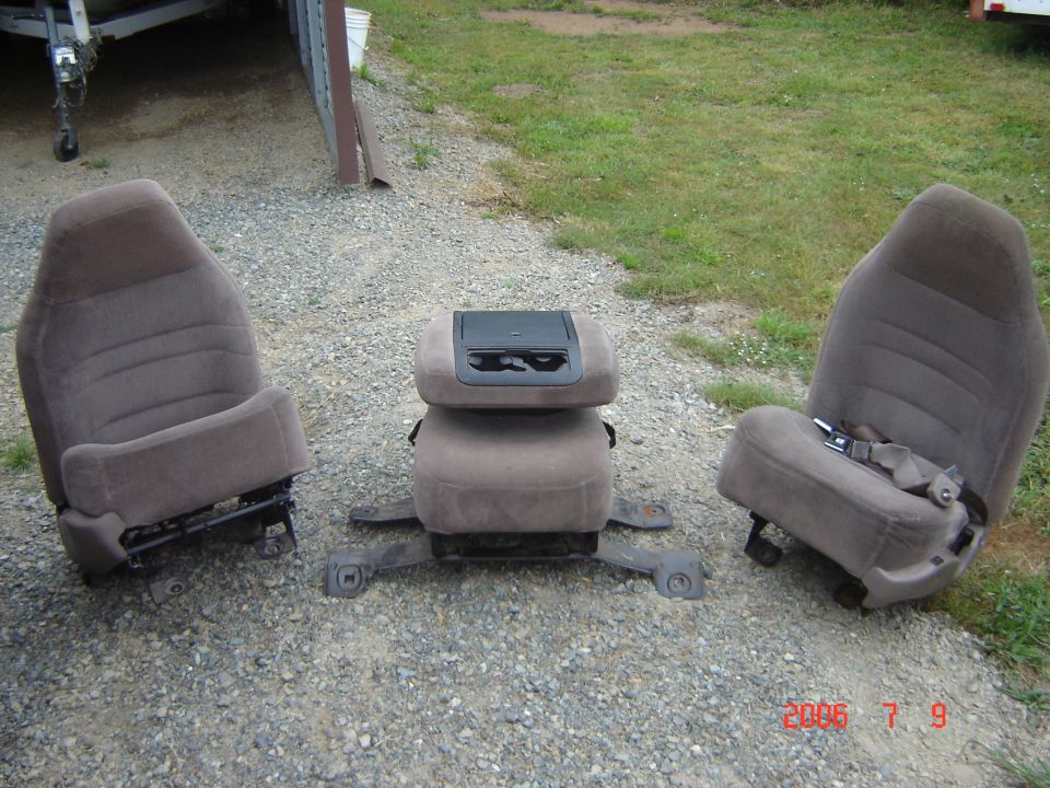 Three removed tan cloth truck seats on gravel, center seat has fold-down cup holder console