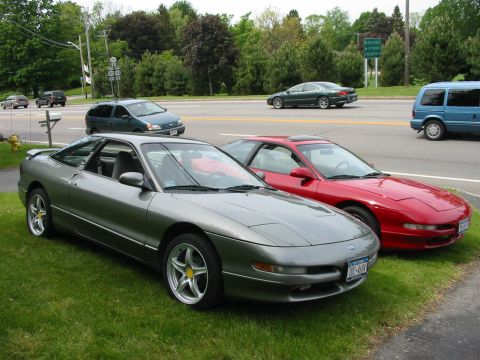 1996 Ford Probe - Two Ford Probes are parked on the grass next to a road with other vehicles in the background.