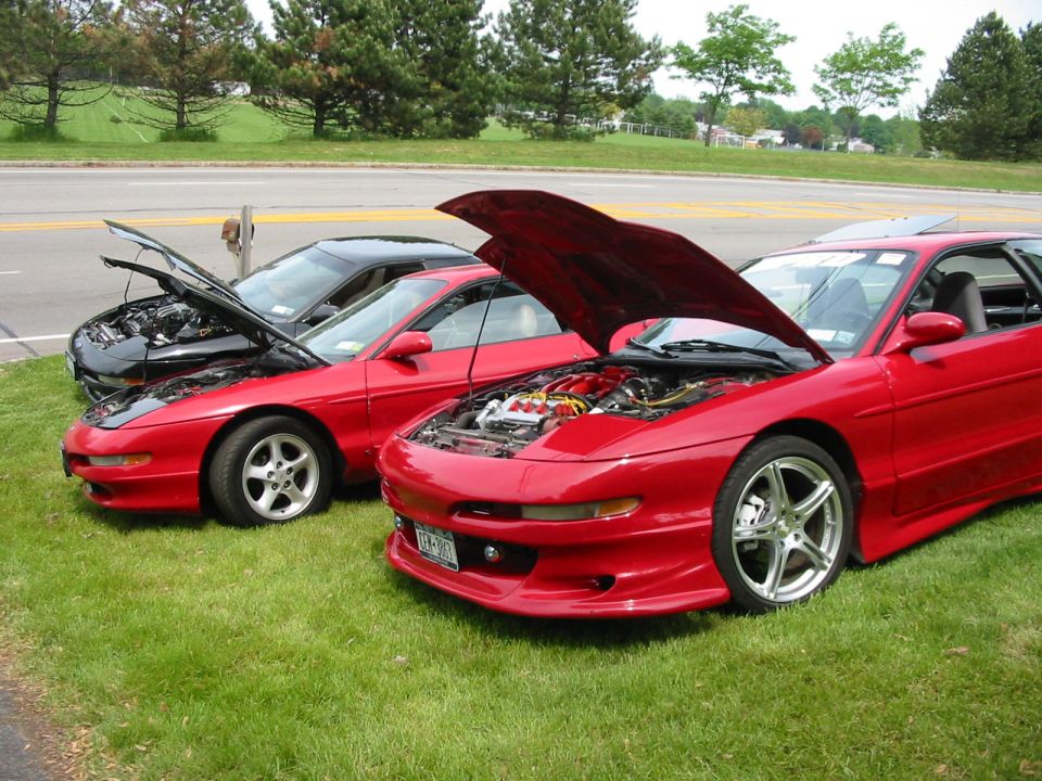 Three Ford Probes parked on grass, hoods open. One is red, one is black, and another is red