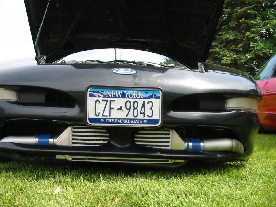Front view of black car with hood open, showing aftermarket intercoolers below the license plate. Another car is visible to the right