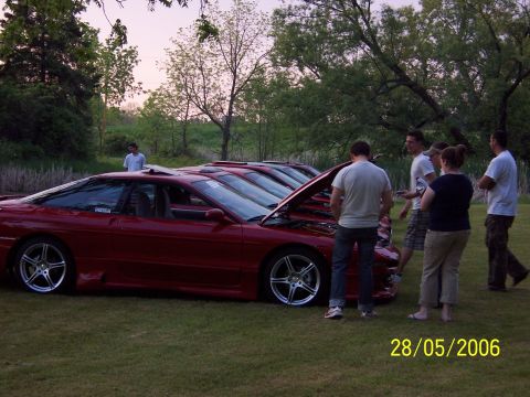1996 Ford Probe - A group of people examine several red coupes lined up on a grassy field, with their hoods open.