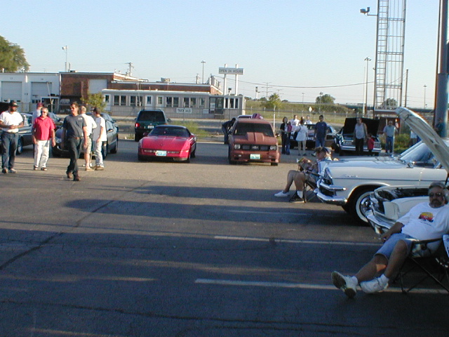 People gathered around cars in a parking lot, some sitting in chairs