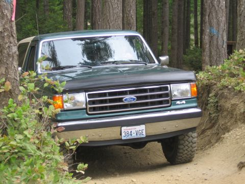 1990 Ford Bronco - A green Ford Bronco navigates a wooded trail, its front grille and 4x4 emblem clearly visible.