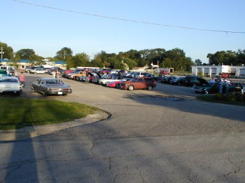 1998 Ford Taurus - A large gathering of vehicles is parked in an outdoor lot, with trees and buildings in the background.