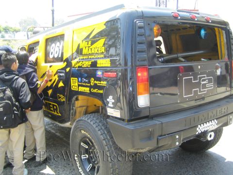 1990 Ford Bronco - Rear three-quarter view of a black Hummer H2 number 861 with sponsor graphics and an H2 logo plate as boys look at it.