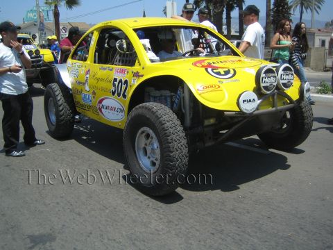 1990 Ford Bronco - Front three-quarter view of a yellow Baja Bug race car number 502 with Hella and PIAA driving lights moving down a street.