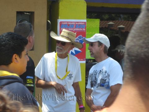 1990 Ford Bronco - Two older men in white shirts and hats stand talking next to a Long Beach Racers Baja 500 event poster on a yellow column.