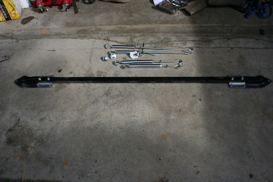 Car parts, including a long black bar and several metal turnbuckles, laid out on a concrete surface